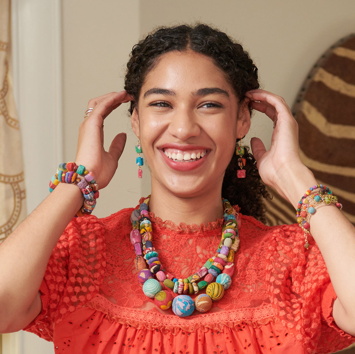 A woman in a red top smiles while touching her colorful, beaded earrings. She is also wearing matching vibrant bracelets and a necklace.