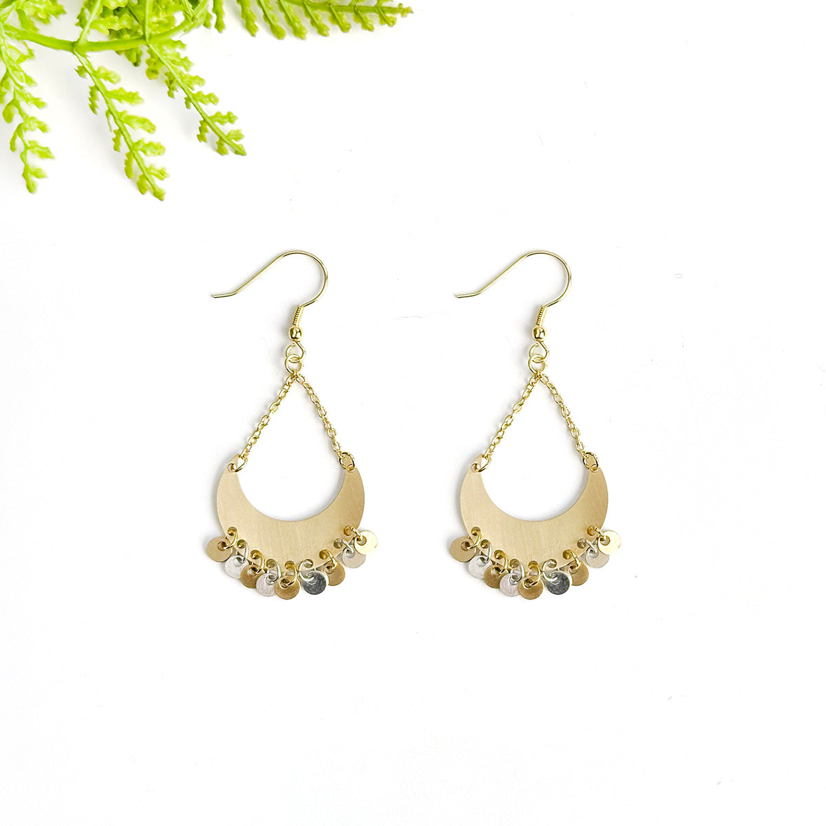 A pair of gold, crescent earrings sits on a white surface with a green leaf in the corner.