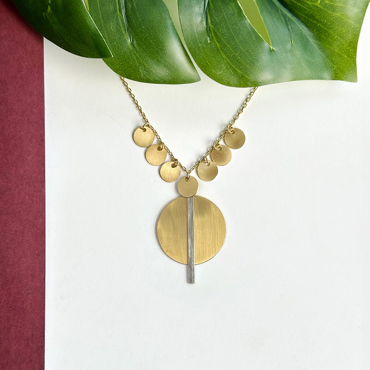 A shiny gold disc necklace is shown against a white backdrop with a large green leaf hanging above it.