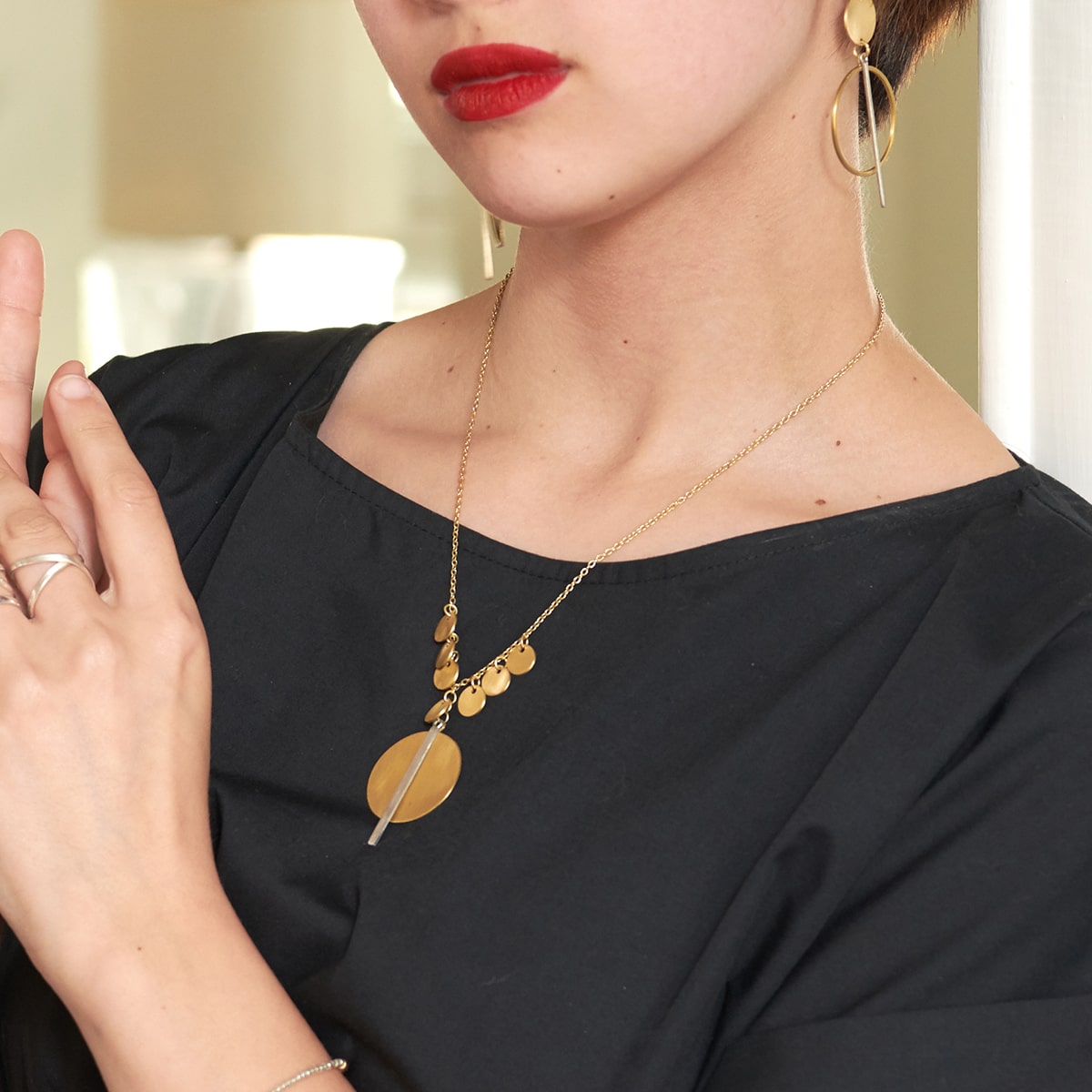 A woman wearing a black dress poses confidently, modeling the gold charm & pendant necklace she is wearing.