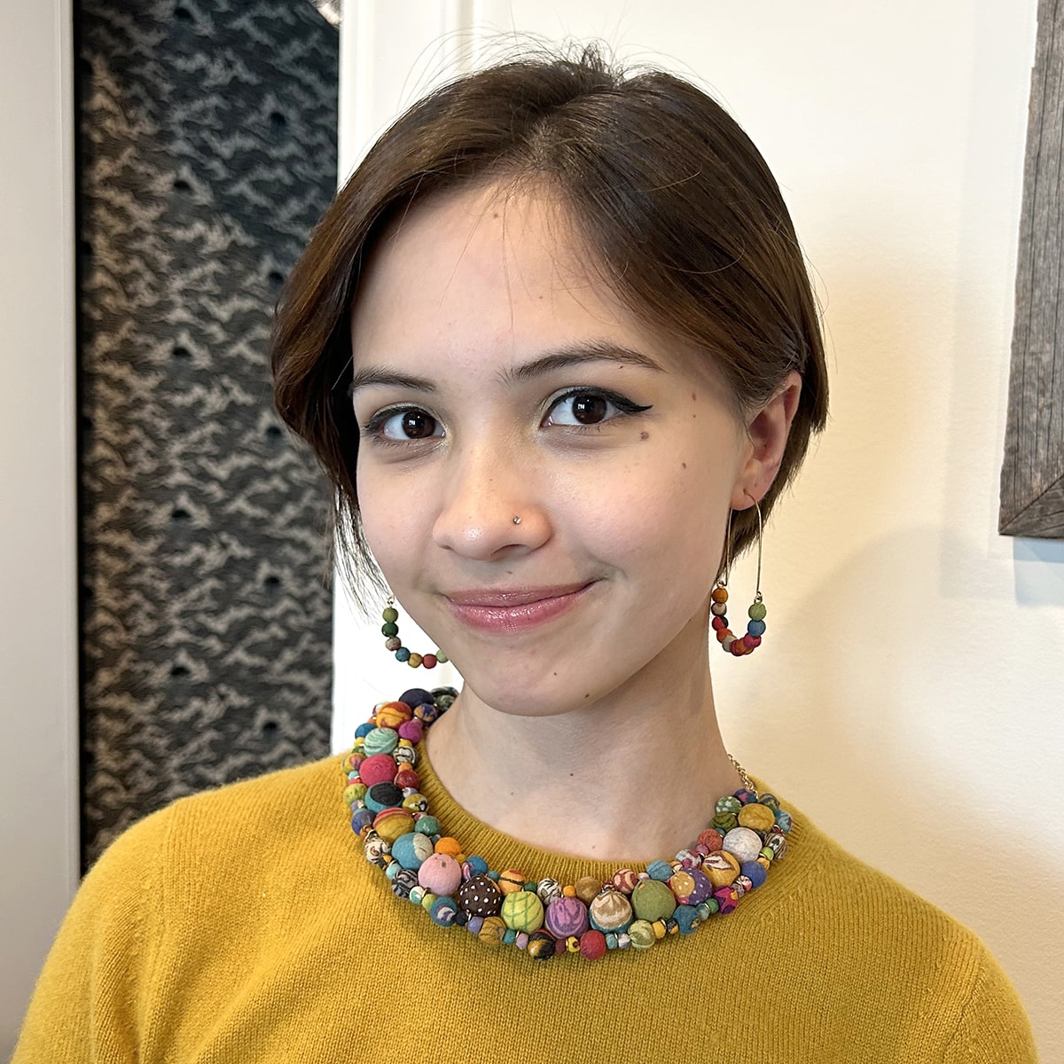 A close up of a woman in a yellow top smiling and modeling her multicolored beaded jewelry.