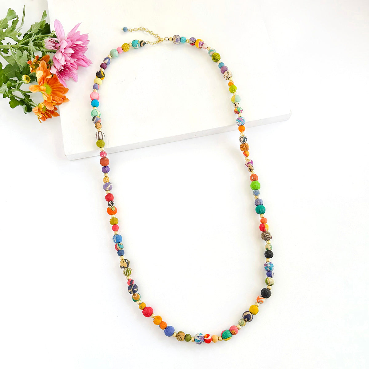 A long beaded bracelet rests on a white backdrop with flowers in the top left corner.
