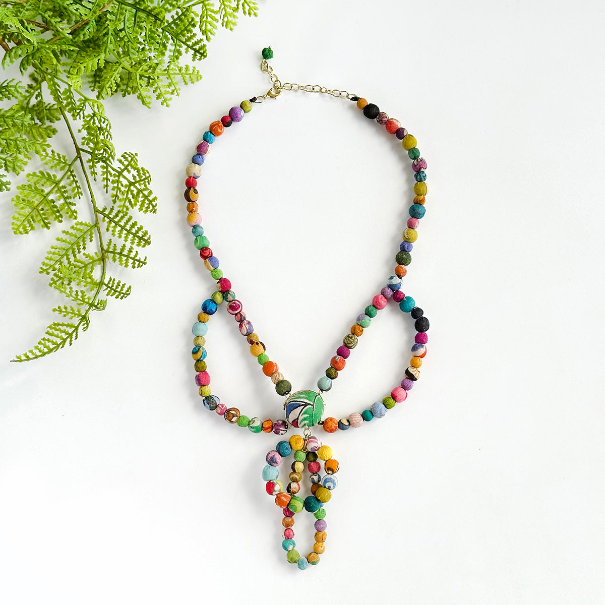 A multicolored beaded necklace rests against a white background, accented by green leaves in the corner.