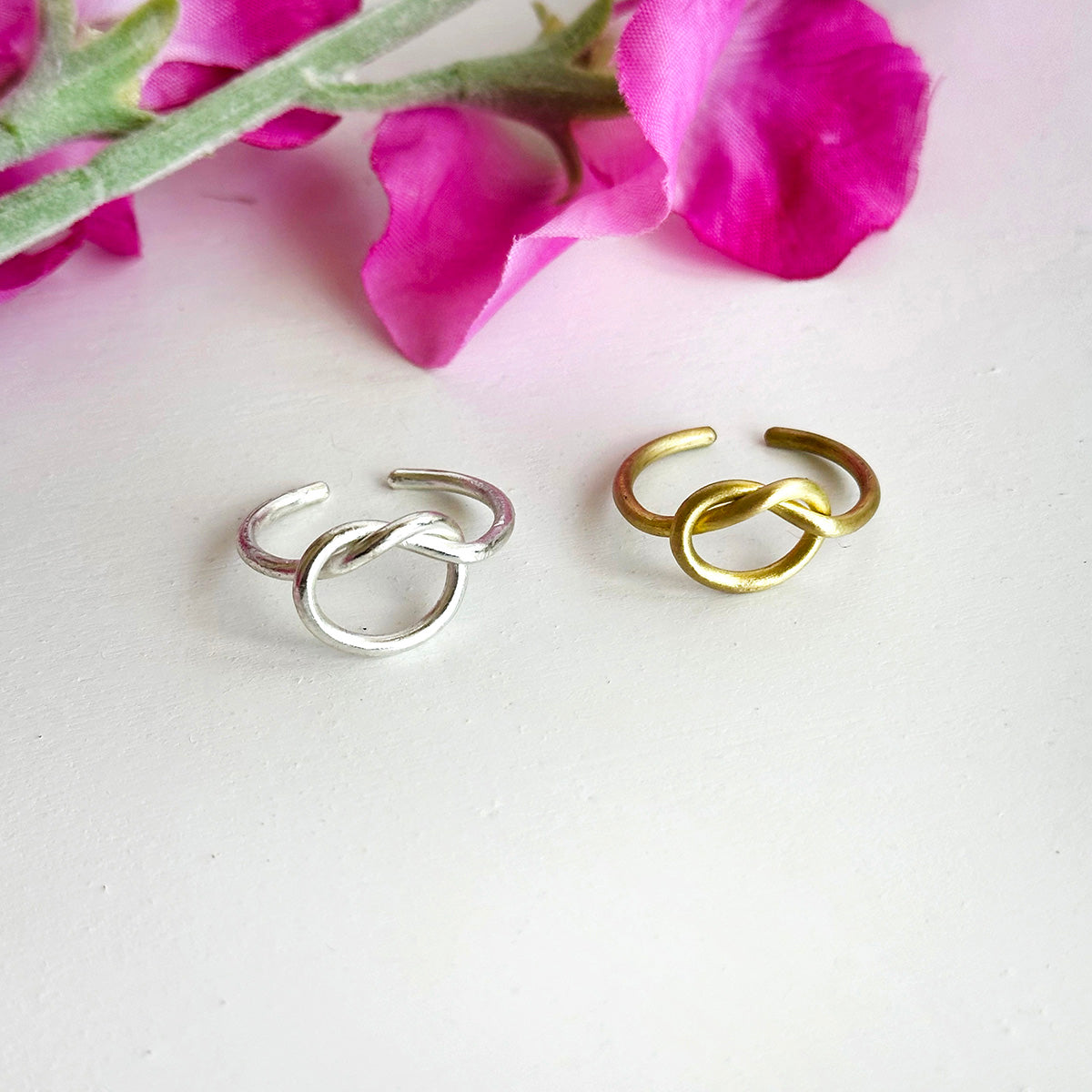 A gold and silver ring, with a center pretzel design, are rest against a white surface with pink flowers in the background.