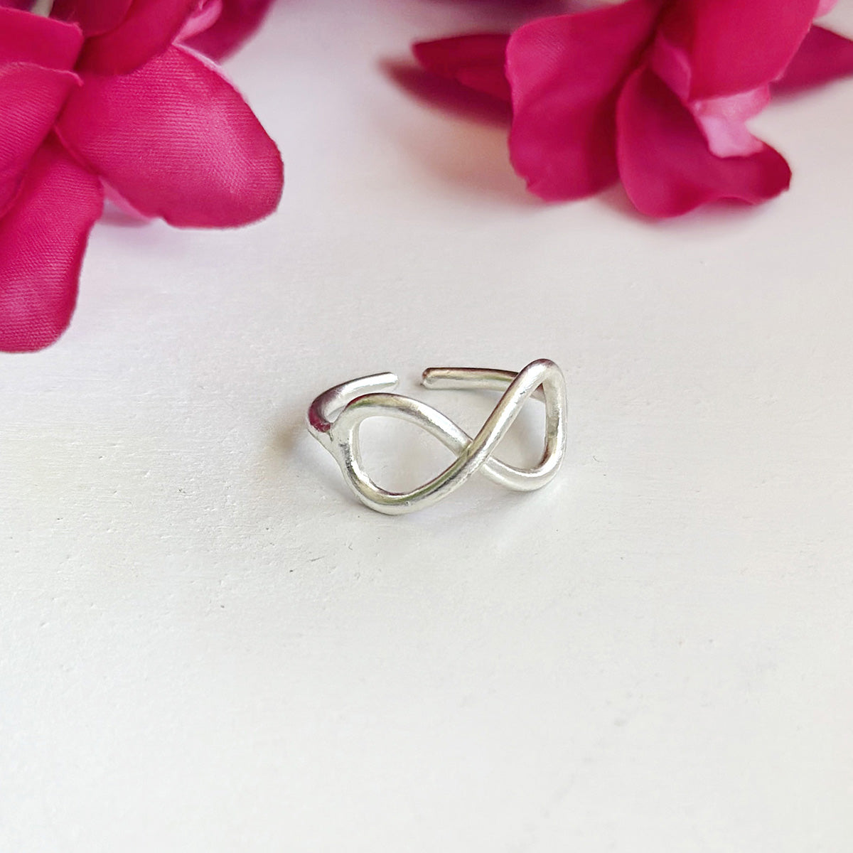 A silver ring with center infinity sign motifs sits against a white surface with a red flower in the background.