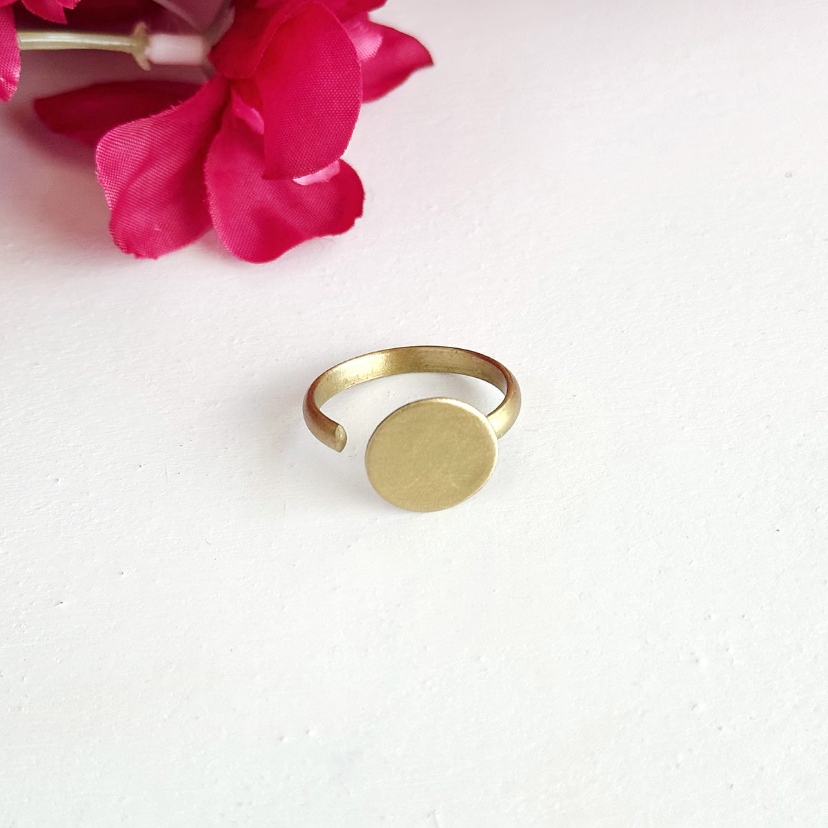 A gold ring with a large circle center motif, is shown against a white background with a pink flower.