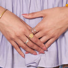 A woman's hands are crossed against her periwinkle shirt, showing her gold and silver rings on her fingers.