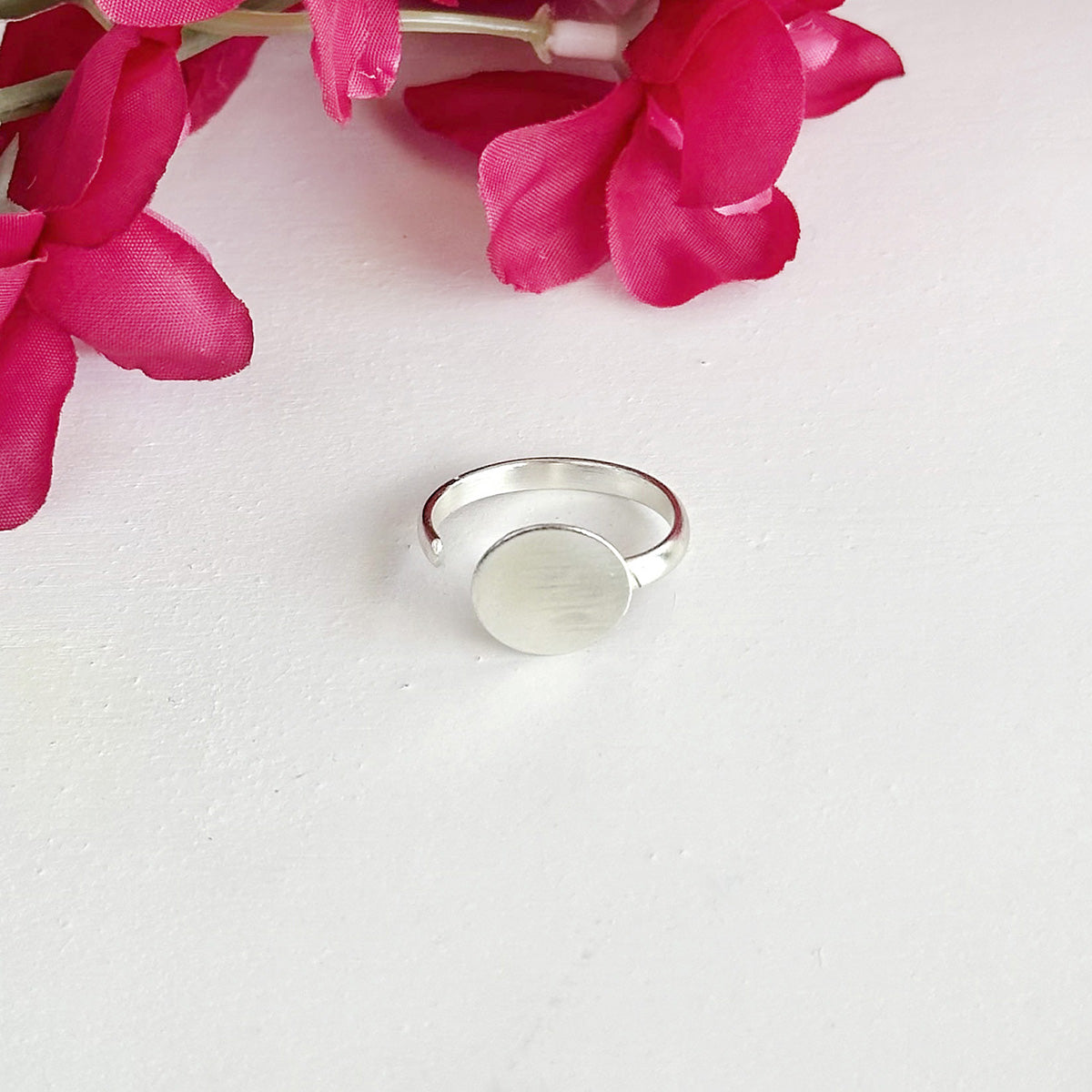 A silver ring with a large circle center motif, is shown against a white background with a pink flower.