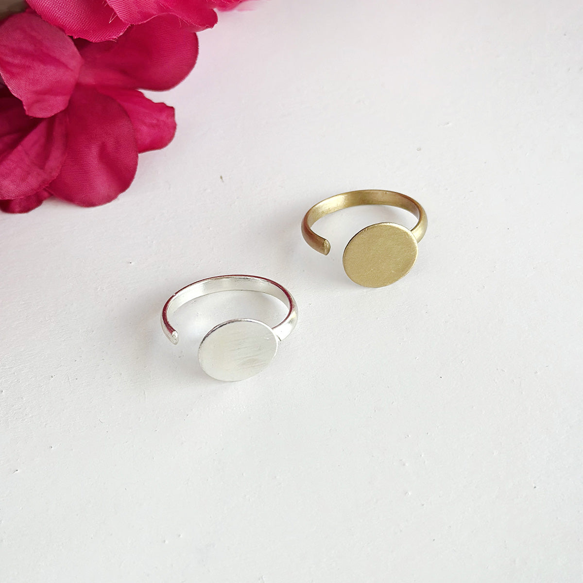 A gold and silver ring, with a large circle center motif, are shown against a white background with a pink flower.