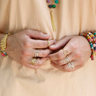A woman in a tan shirt clasps her hands together, showing her many gold and silver rings on her fingers.