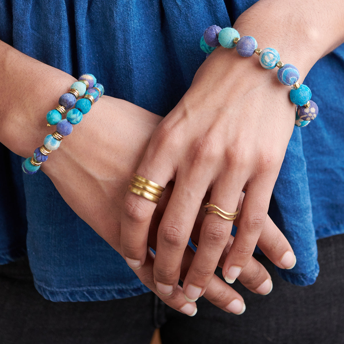A woman in an blue shirt holds her hands together, displaying her blue beaded jewelry and two metallic rings.