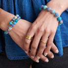 A woman in an blue shirt holds her hands together, displaying her blue beaded jewelry and two metallic rings.
