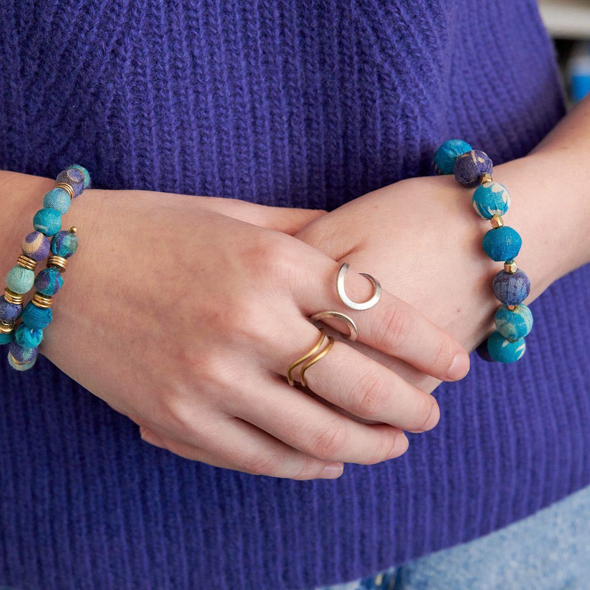 A woman in an indigo sweater holds her hands together, displaying her blue beaded jewelry and two metallic rings.