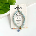 A blue beaded bracelet tied to a card that reads "Inspire" against a white background with a green leaf to the left.