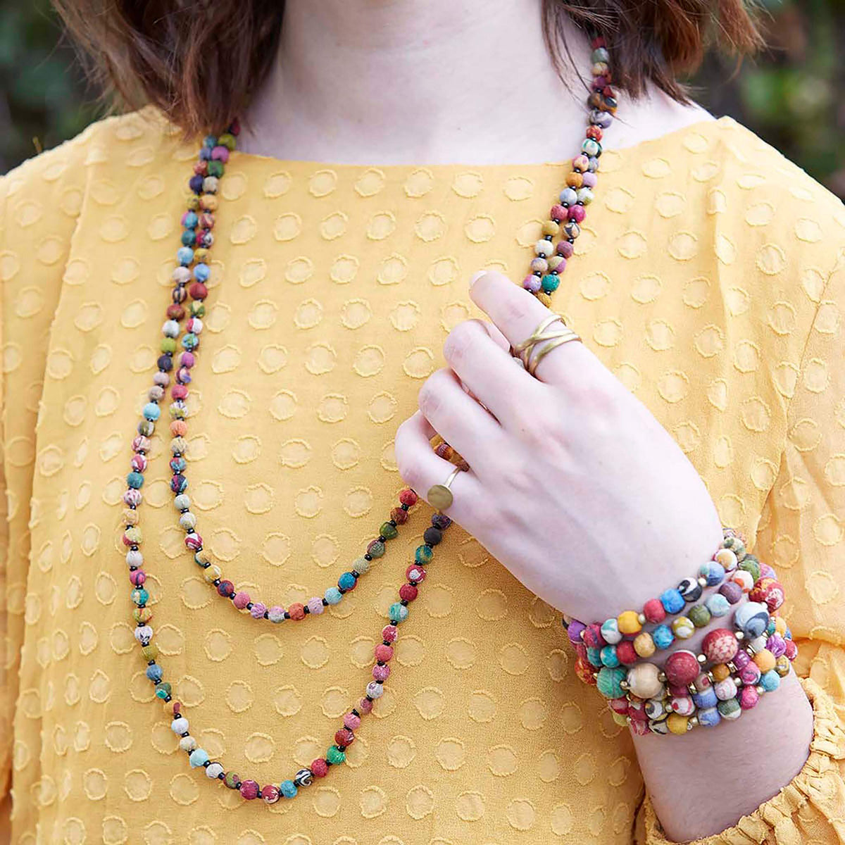A women in yellow touches her multicolor, long beaded necklace, highlighting a stack of coordinating beaded bracelets.