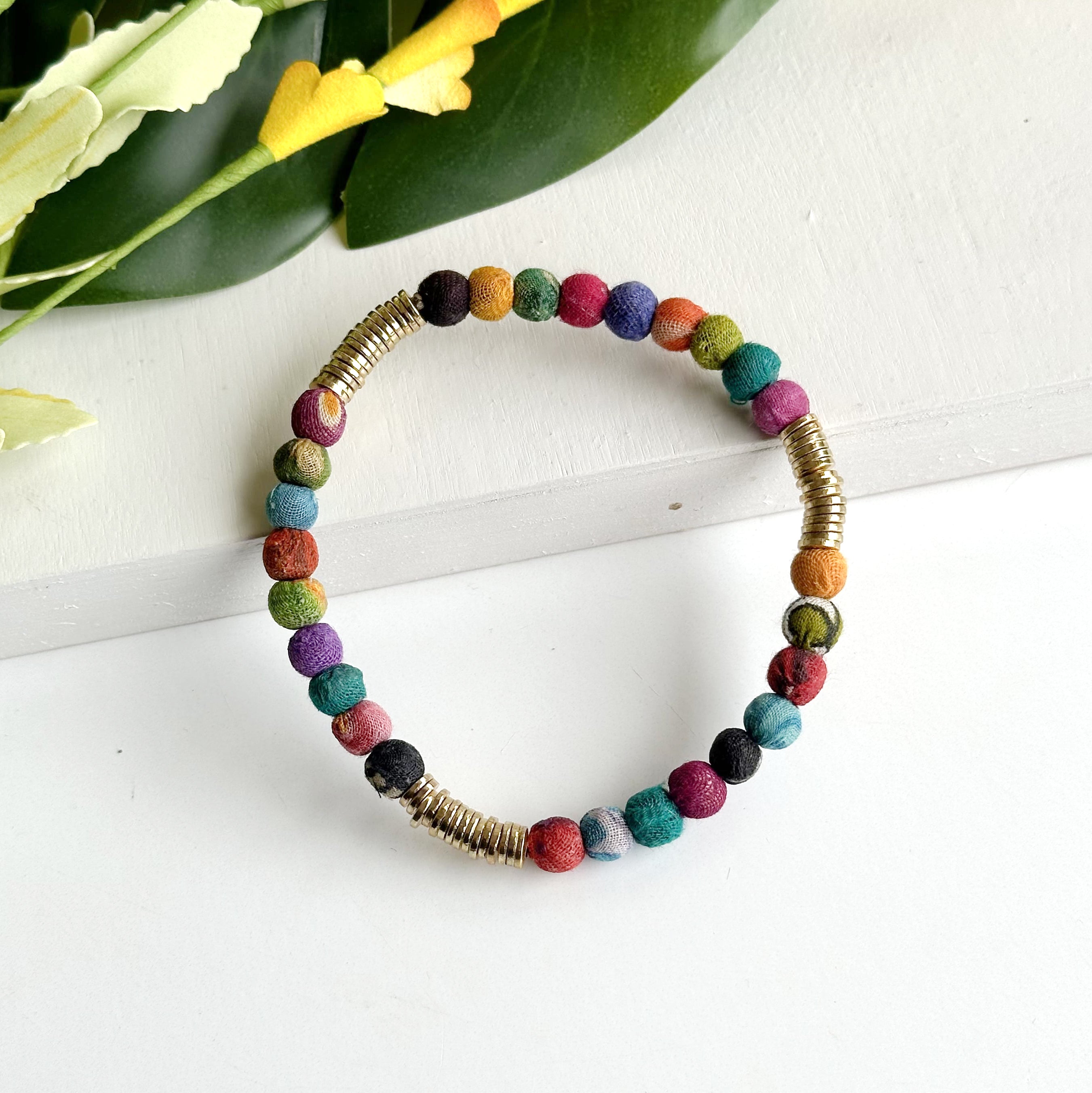 A multicolor beaded bracelet rests on a white background with a green and yellow flower in the top left corner.