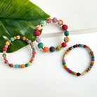 Three multicolor beaded bracelets rest on a white surface with a large green leaf in the top left.