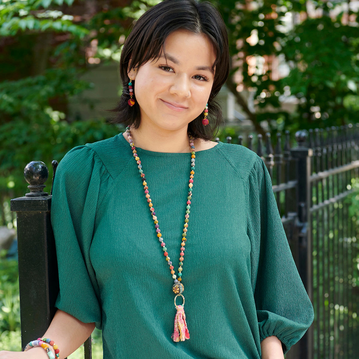 A woman in a teal shirt and multicolor, beaded jewelry leans against a fence.
