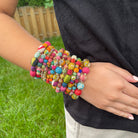A womans hand rests on her hip, showing off her wrist with a stack of twelve multicolor bracelets.