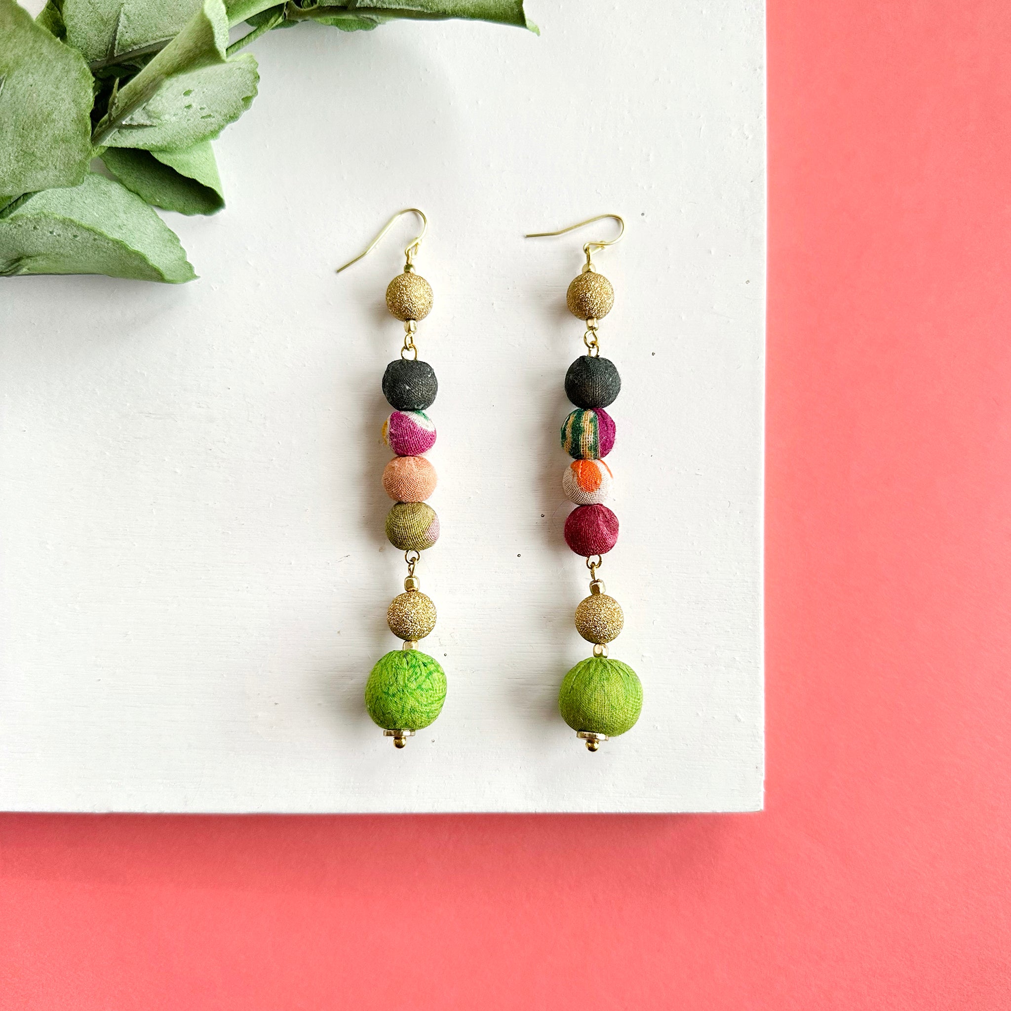 A pair of earrings is shown against a white and pink background.Two shimmering gold beads sit atop a stack of 4 colorful Kantha beads and a larger anchor bead.