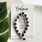 A black and white beaded bracelet tied to a card that reads "Balance" against a white background with a green leaf to the left.