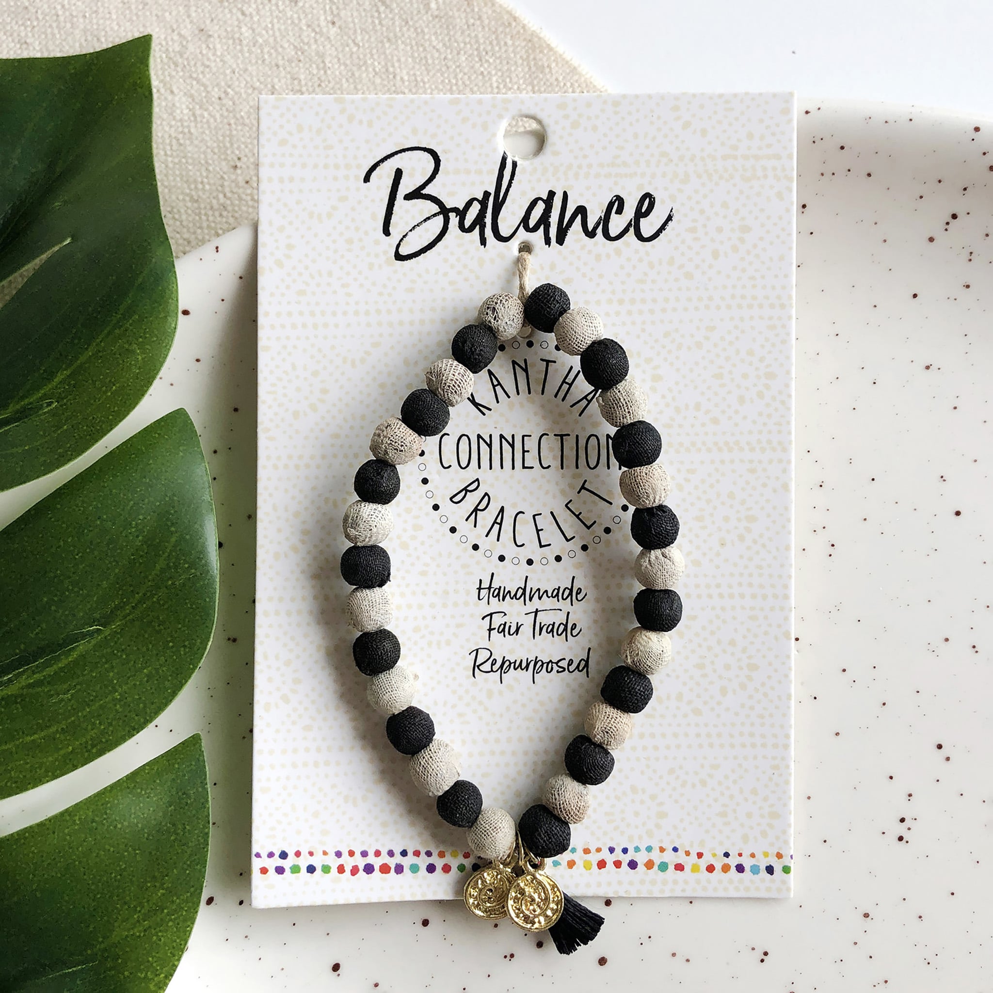 A black and white beaded bracelet tied to a card that reads "Balance" against a white background with a green leaf to the left.