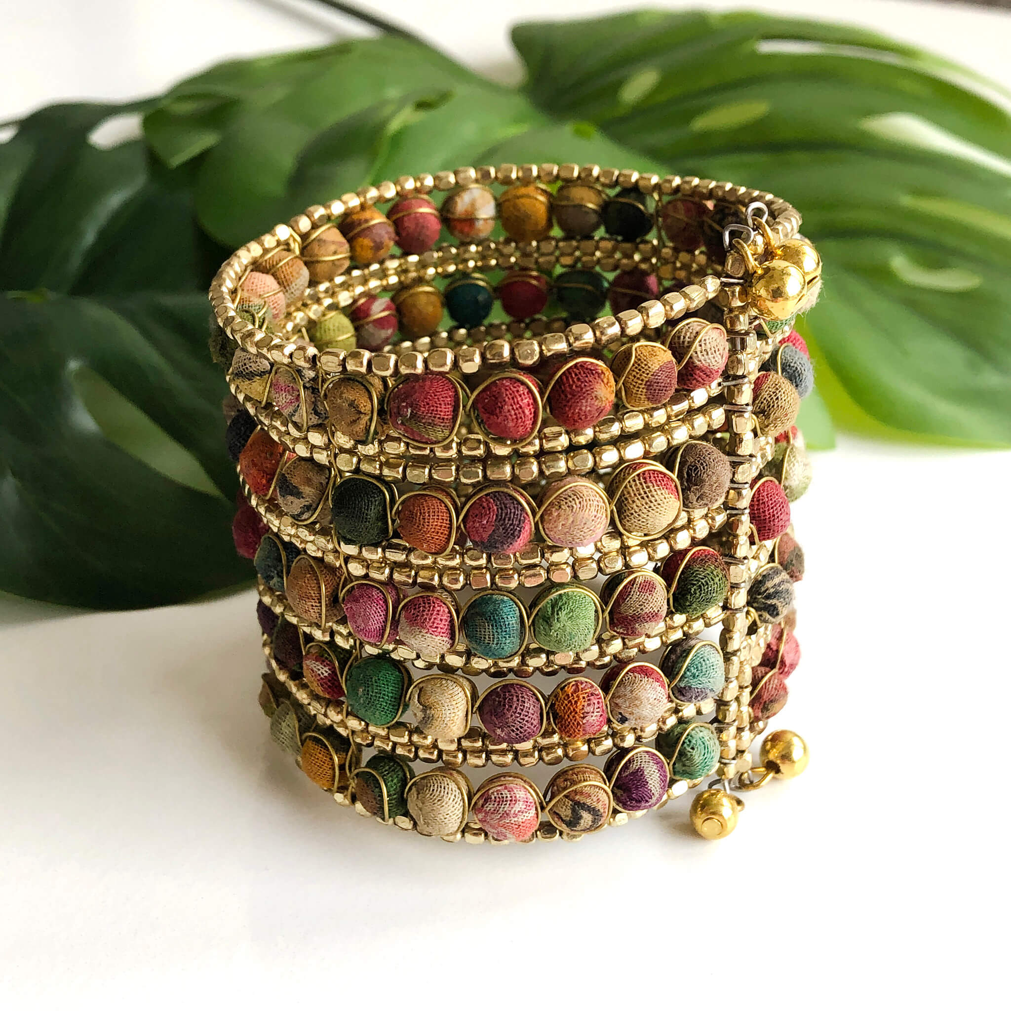 A large cuff bracelet made from many small textile-covered beads and tiny gold beads against a white background and green leaves.