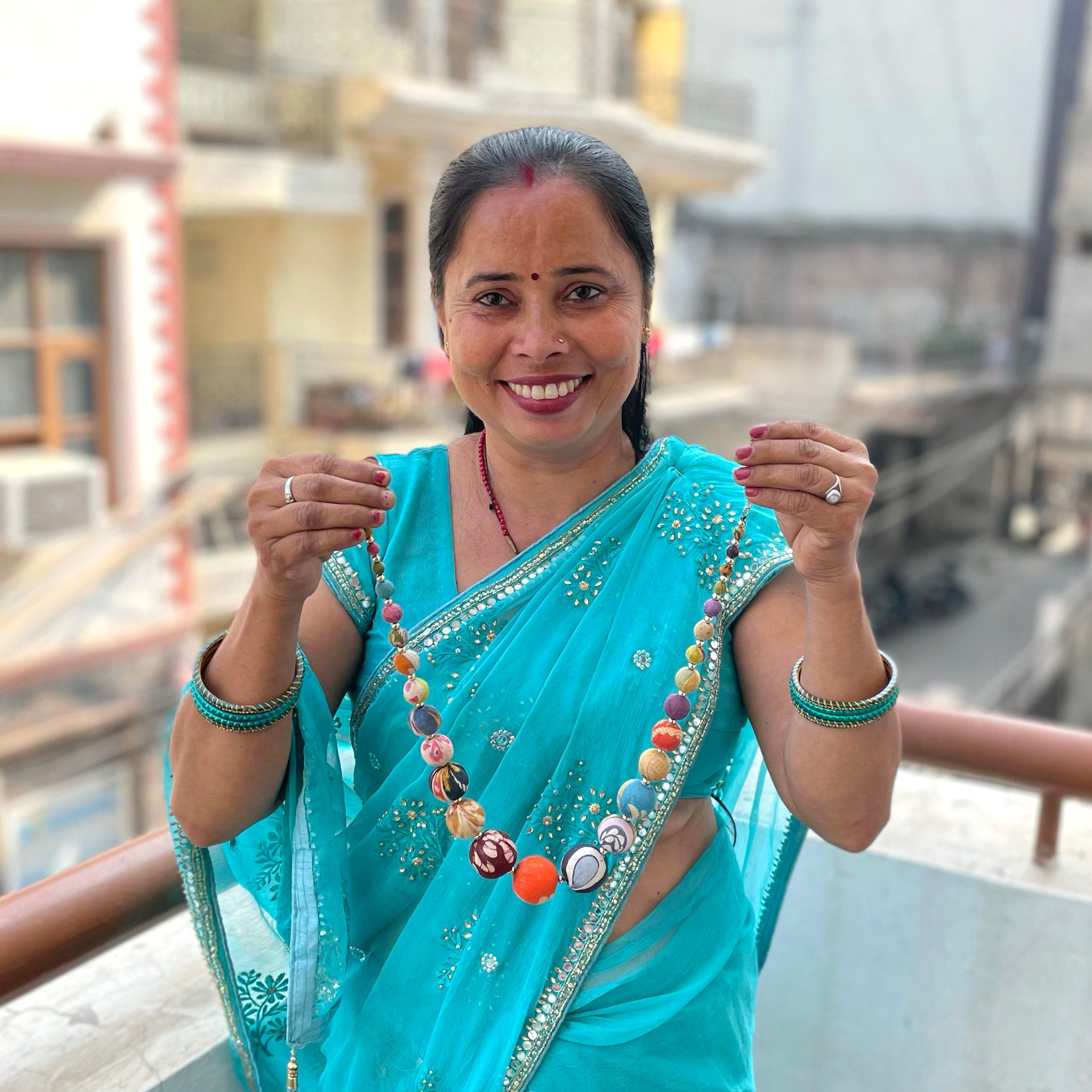 A woman in a blue Sari holds up a multicolor, beaded necklace, and smiles.
