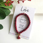 A red beaded bracelet tied to a card that reads "Love" against a white background with a green leaf to the left.