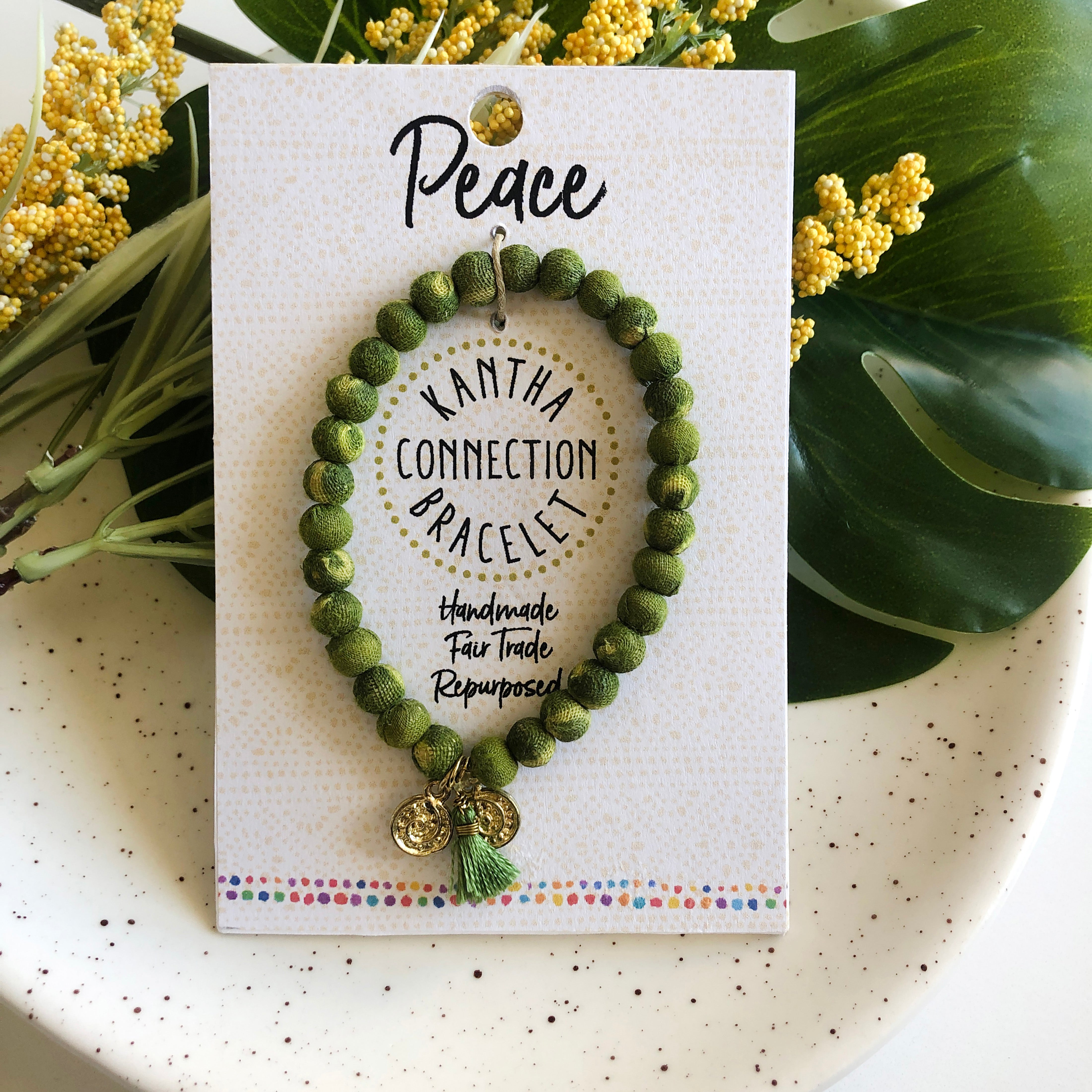 A green beaded bracelet tied to a card that reads "Peace" against a white background with a green leaf to the left.