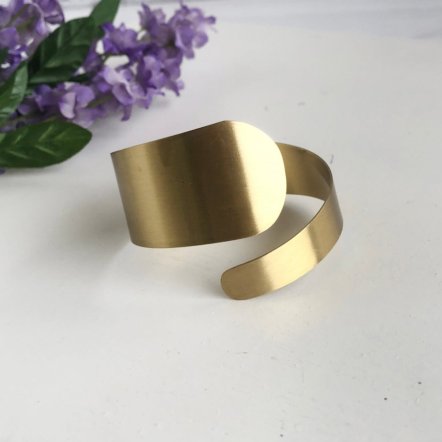 A gold spiral/swirl-inspired cuff bracelet is seen with a purple flower in the background.