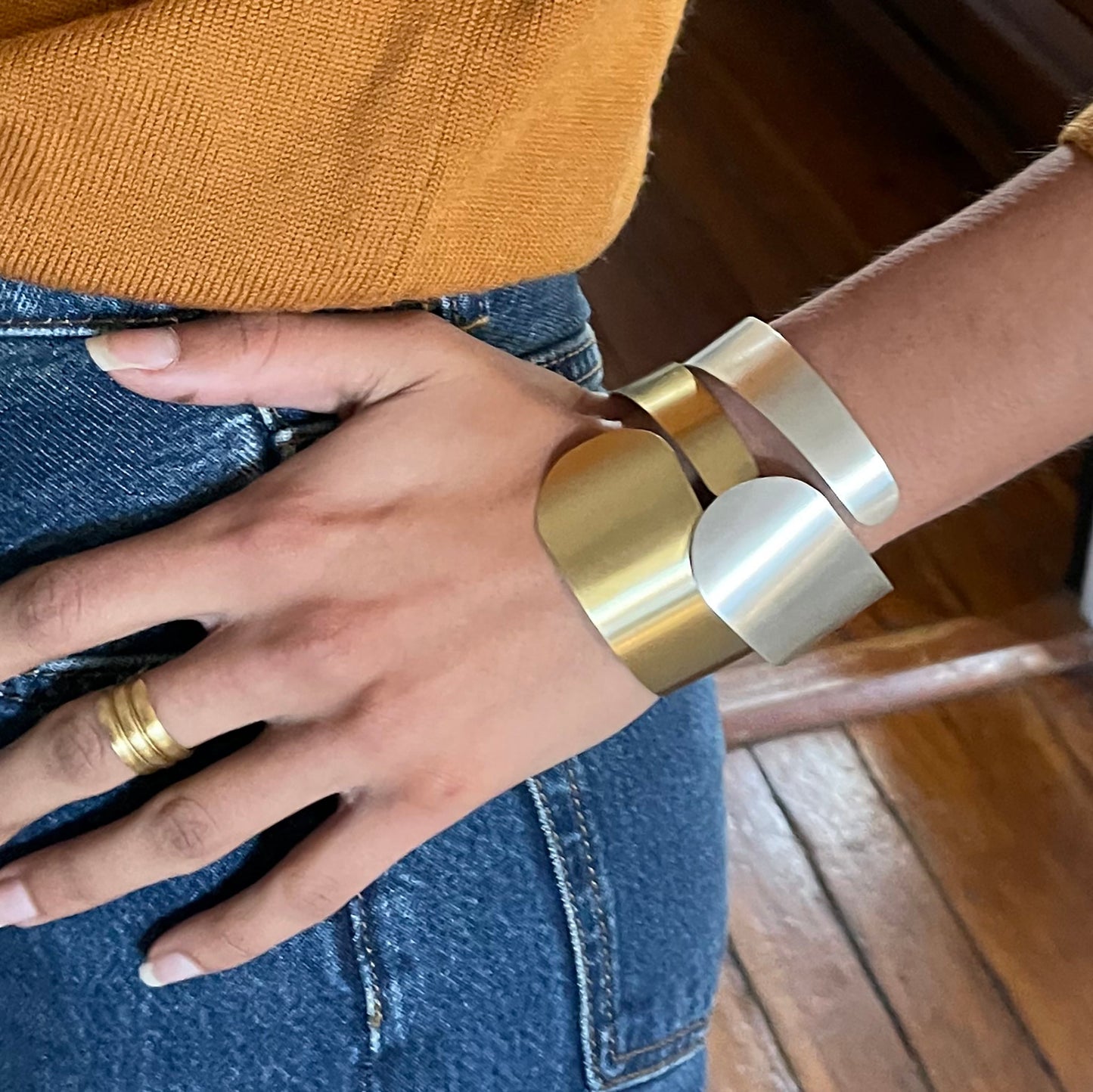 A close up image of a a woman's hand resting on her hip, adorned with two Wraparound Cuff bracelets, one in gold and one in silver.