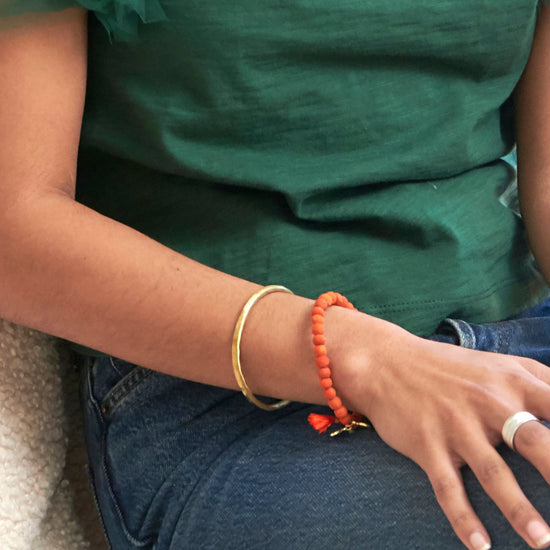 A woman's hand rests on her leg, highlighting a gold cuff bracelet and an orange beaded bracelet.