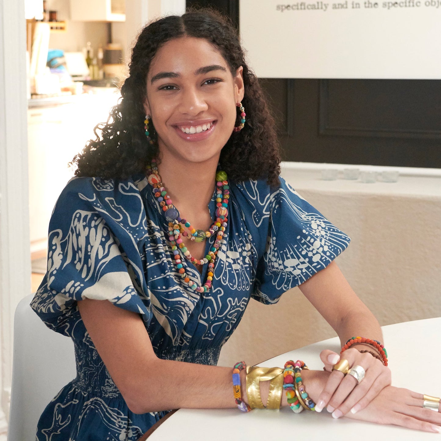 A woman smiles at us while adorned with colorful beaded jewelry and a large gold metallic cuff.