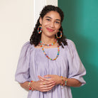 A young woman in a lavender shirt smiles while showing off her multicolor, beaded jewelry.