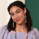 A young woman in a purple shirt smiles while showing off her multicolor, beaded jewelry.