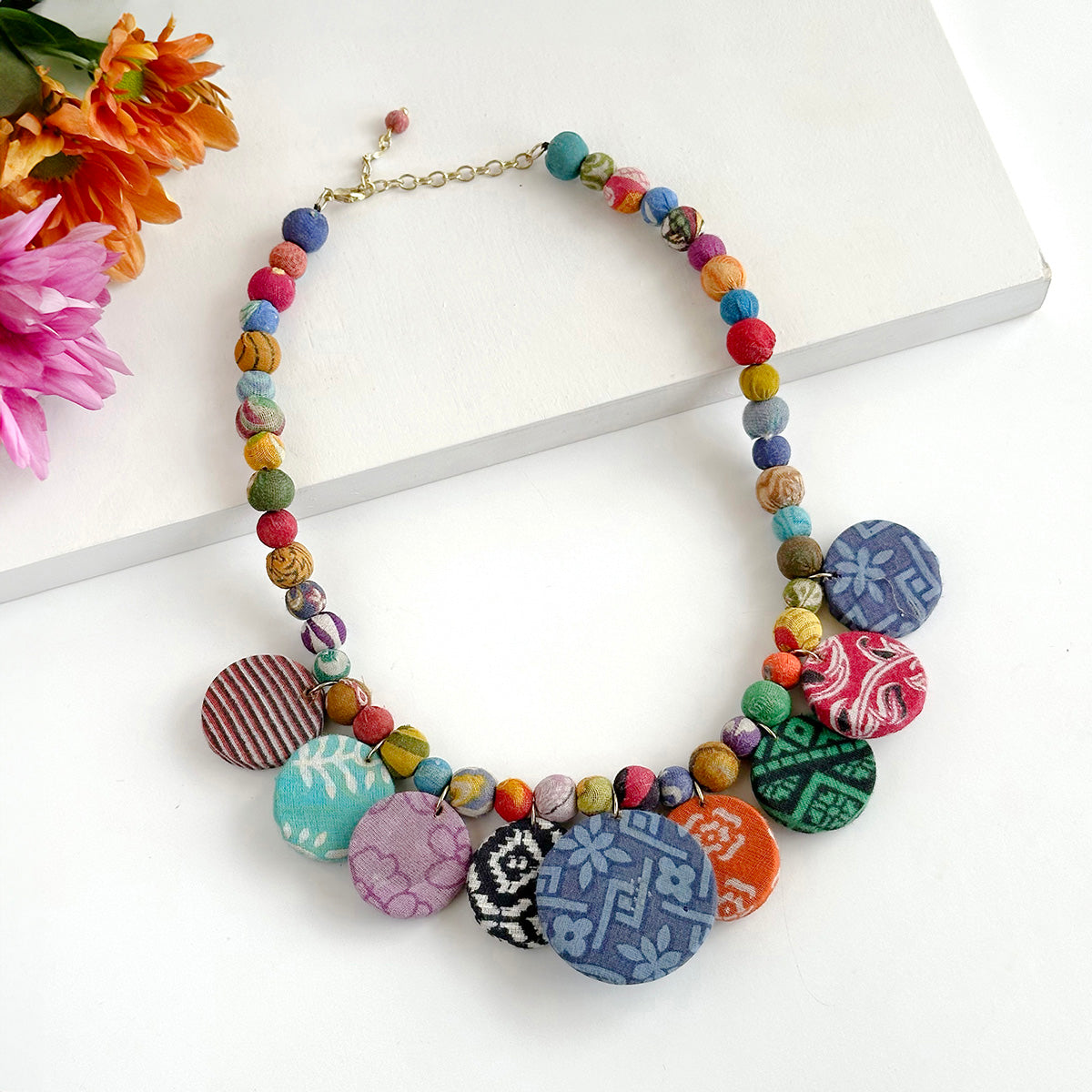 A multicolored beaded statement necklace rests against a white board, accented by a pink and orange flowers in the corner.