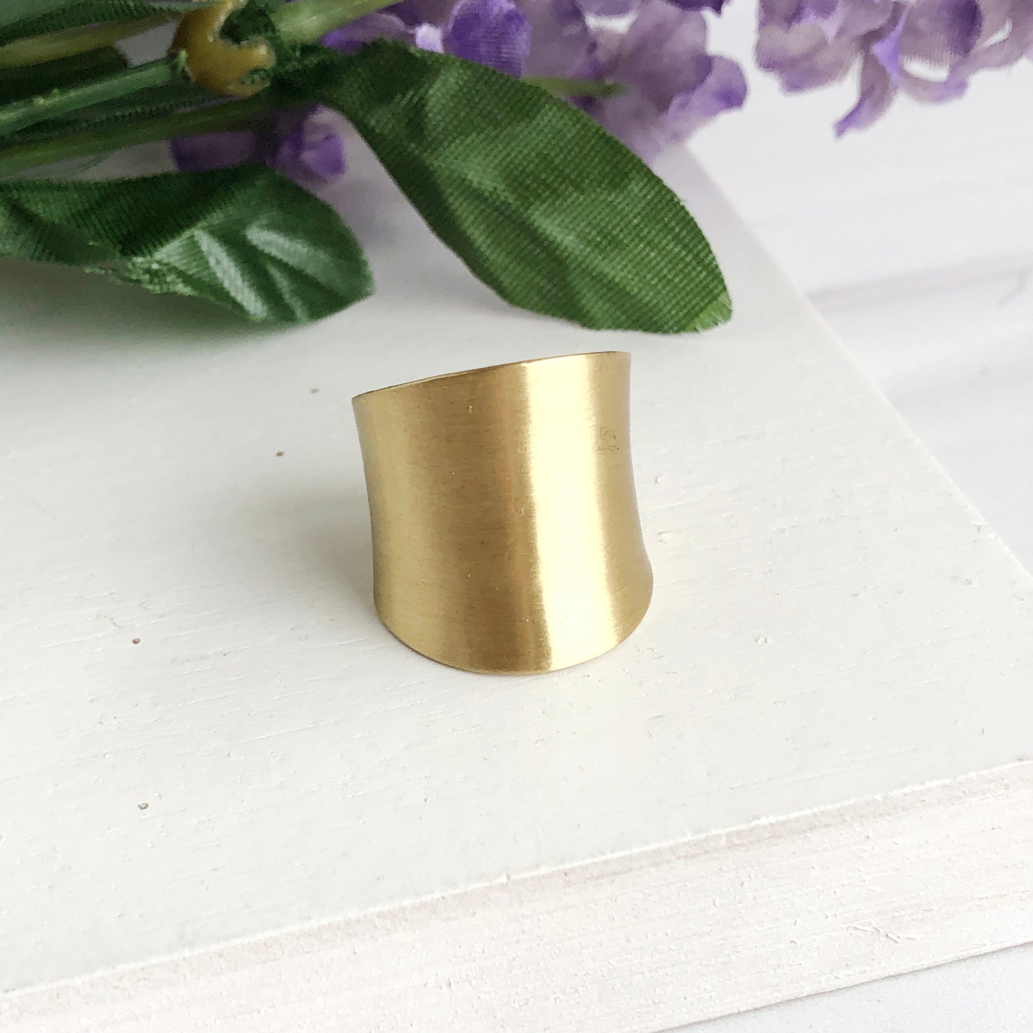 A small, but tall gold ring is placed against green & purple flowers.