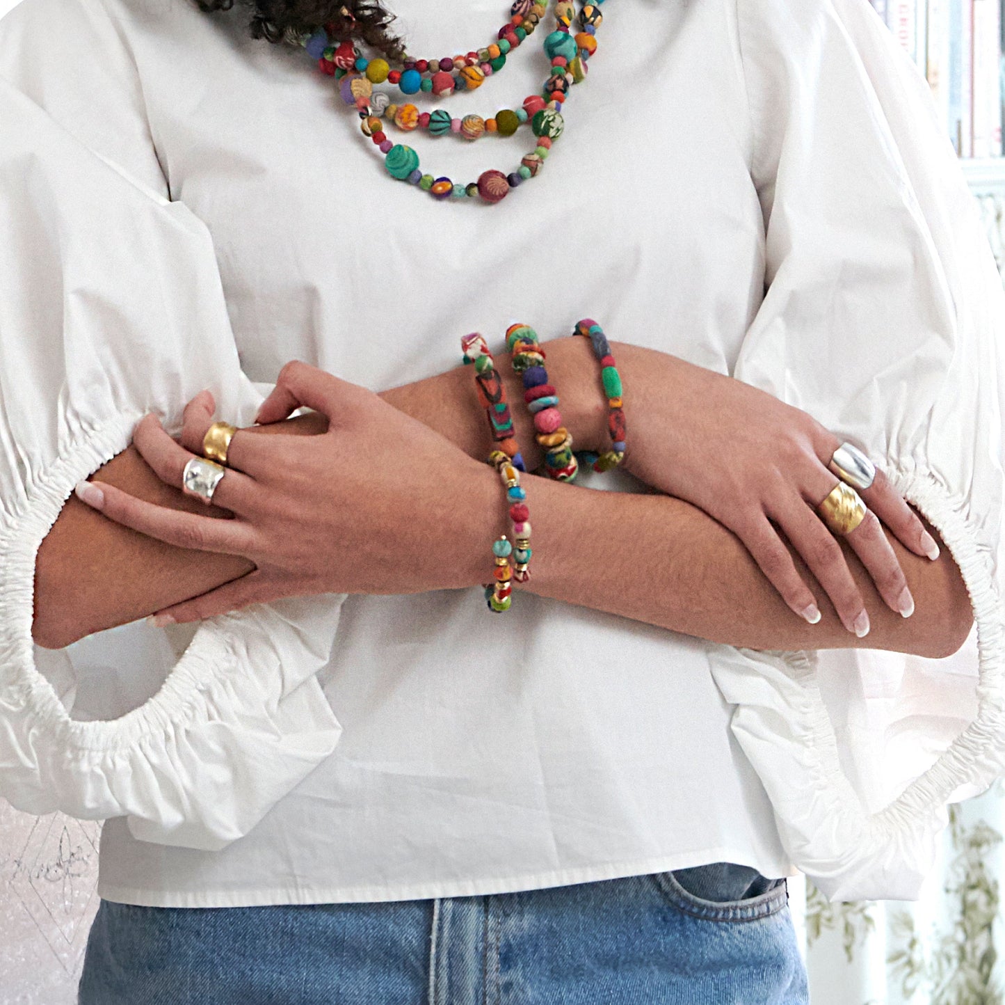 A woman crosses her arms across her chest, showing a stack of colorful beaded bracelets and metallic rings.
