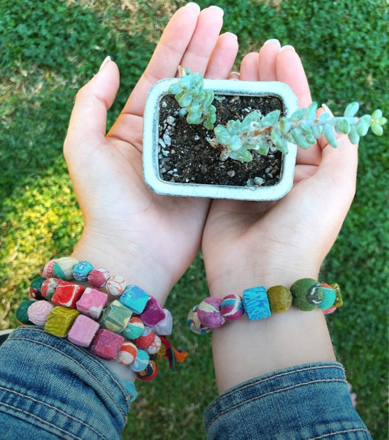 Two hands hold a small potted plant while wearing colorful beaded Kantha bracelets