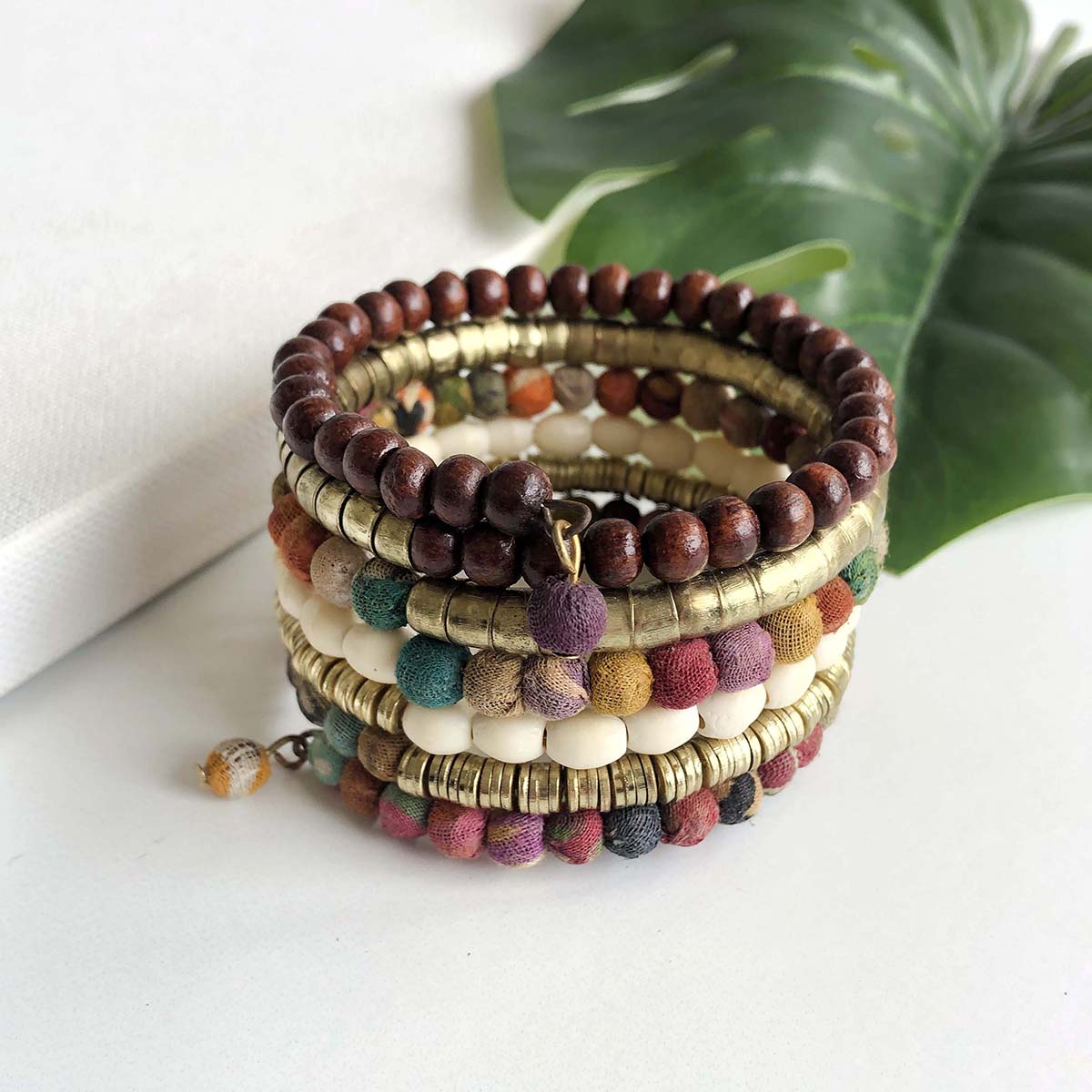 A colorful wrap-style cuff bracelet is shown against a white surface with a dark green leaf in the background.