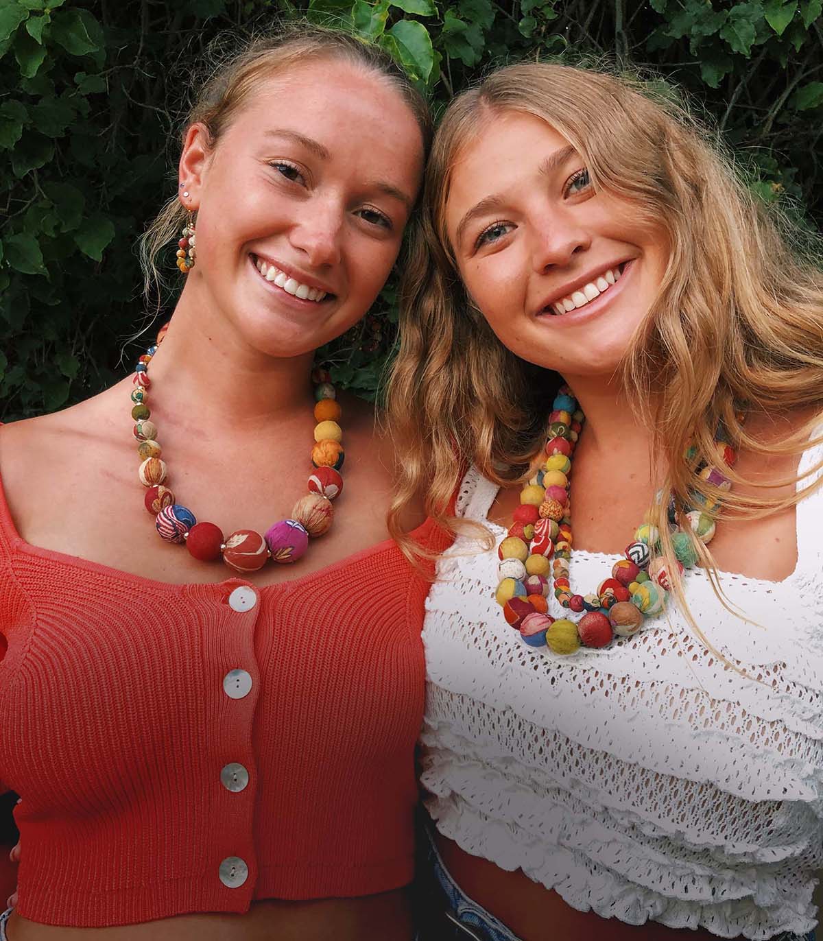 Two women smile at the camera while modeling colorful beaded jewelry.