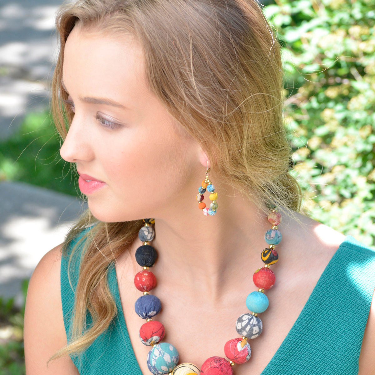 A woman in a teal shirt and multicolor, beaded jewelry looks off to the left.