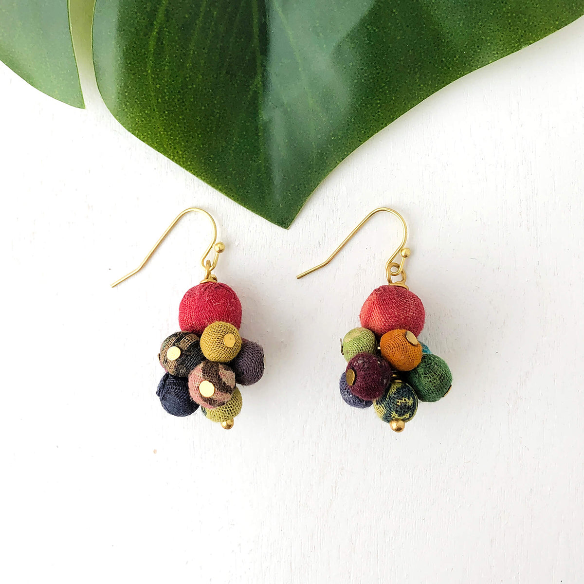 A pair of small, multicolor, multi-bead earrings against a white background with a large green leaf along the top.