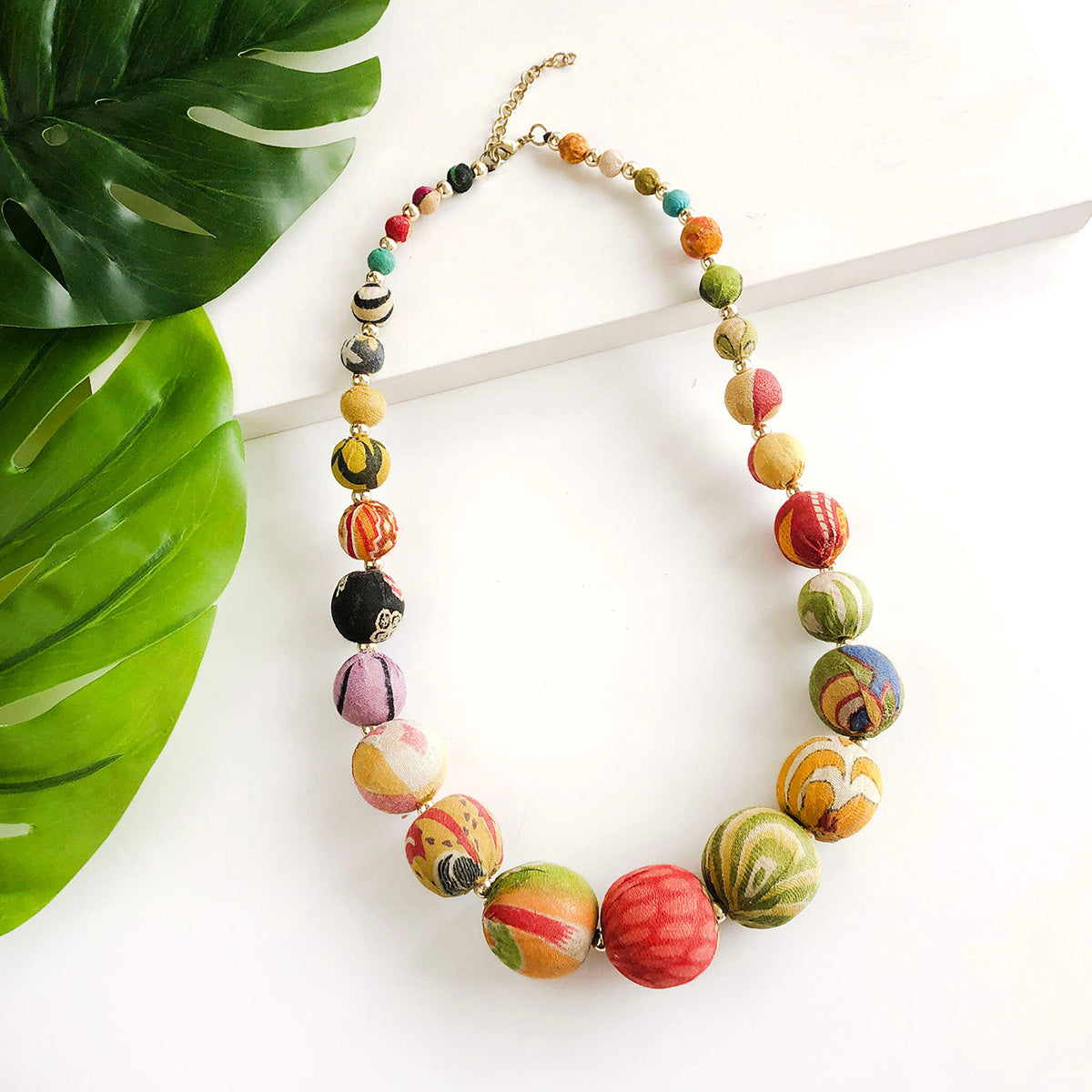 A colorful, beaded necklace against a white background with green leaves in the corner.