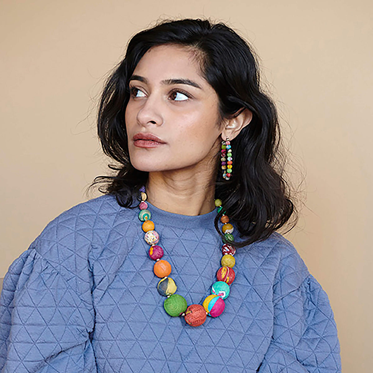 A woman in a blue shirt looks to the side and is adorned with multicolor, beaded jewelry.