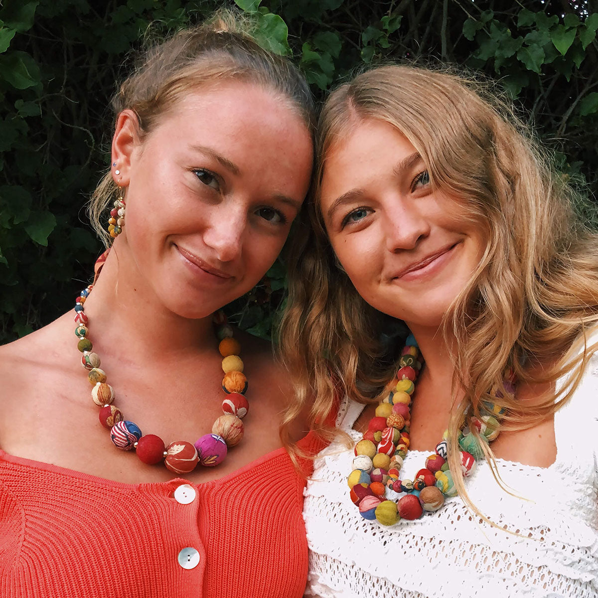 Two young women smile at while wearing colorful Kantha necklaces.