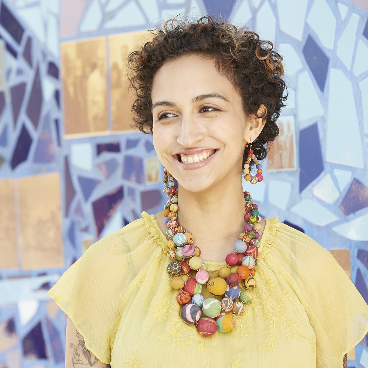 A woman wearing a yellow shirt smiles while wearing a large, layered multi colored beaded necklace.