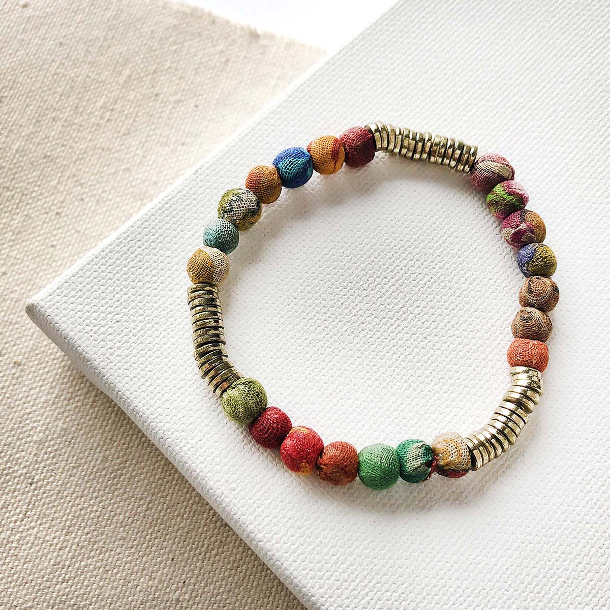 A single strand bracelet of multicolor and gold beads rests on a white block against a tan background.