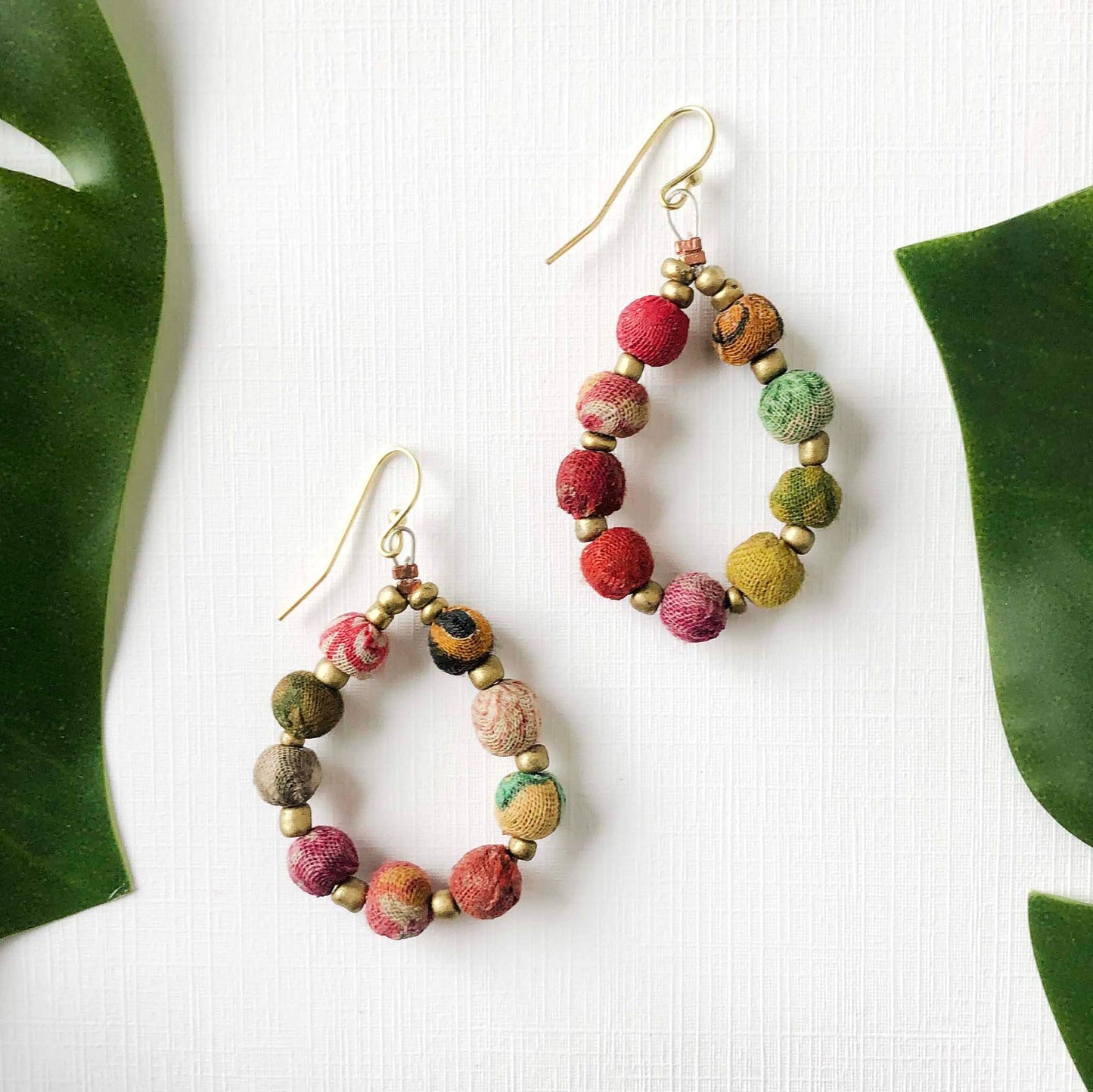A pair of colorful textile-wrapped beaded earrings are flanked by green leaves.